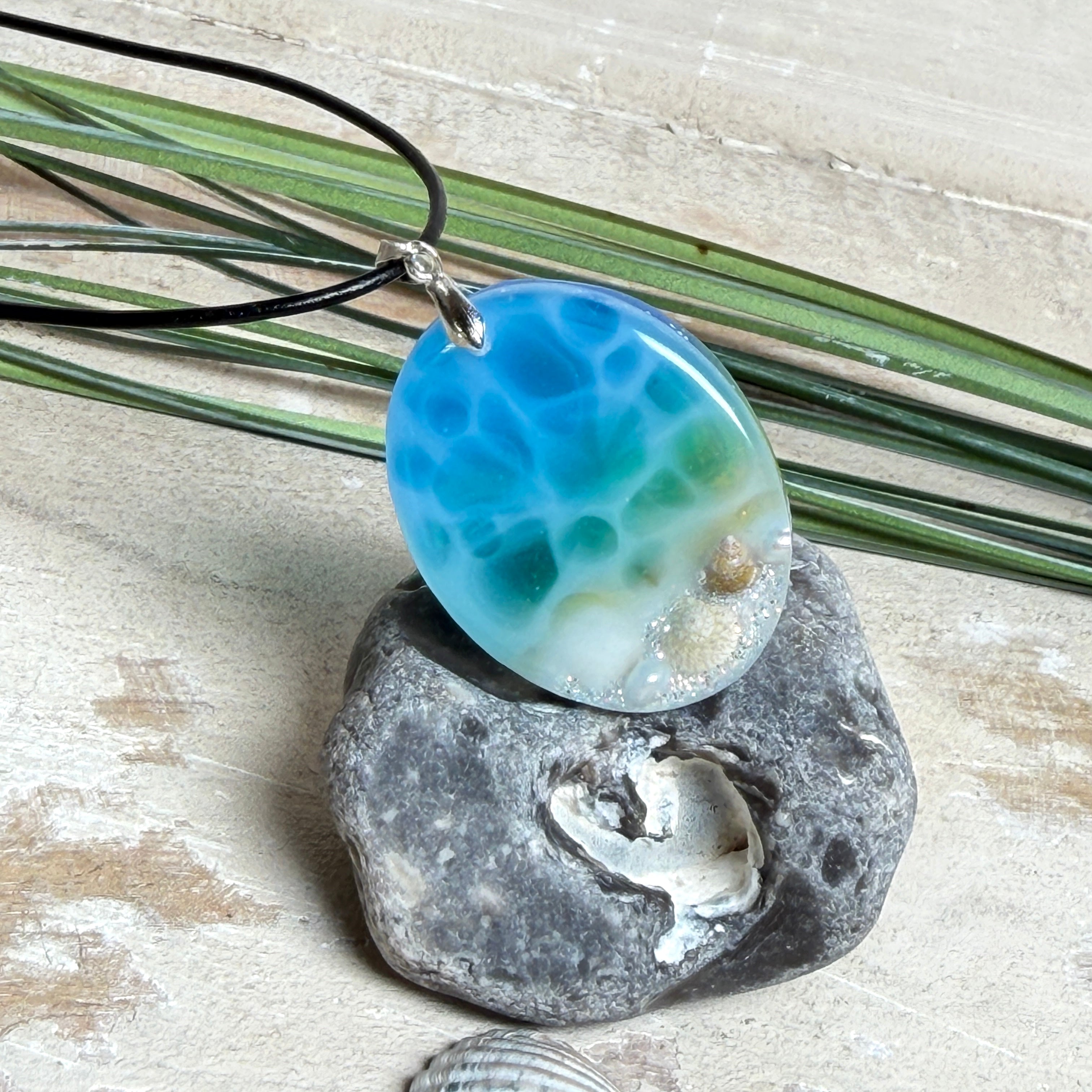 Ovale Harz-Kette „Ocean Oval“ mit ca. 4 x 3 cm großem Anhänger in Blau-Grün-Weiß-Verlauf. Am unteren Rand sind kleine Muscheln, Sandstruktur und feiner Glitzer eingegossen, sodass der Eindruck eines sanften Strandes im klaren Wasser entsteht. Der Anhänger besitzt eine Edelstahl-Schlaufe und hängt an einer schwarzen Lederkette mit Edelstahl-Karabiner und Verlängerung. Maritimer Unikat-Schmuck für Meeresliebhaberinnen.