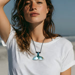 Walflossen-Kette aus Holz und türkis-weißem Harz an schwarzem Lederband, getragen von einer Frau im weißen T-Shirt am Strand – handgefertigter Unisex-Oceanschmuck von ZeitlosSchmuckDesign für Meerliebhaber und Freiheitsgefühle.
