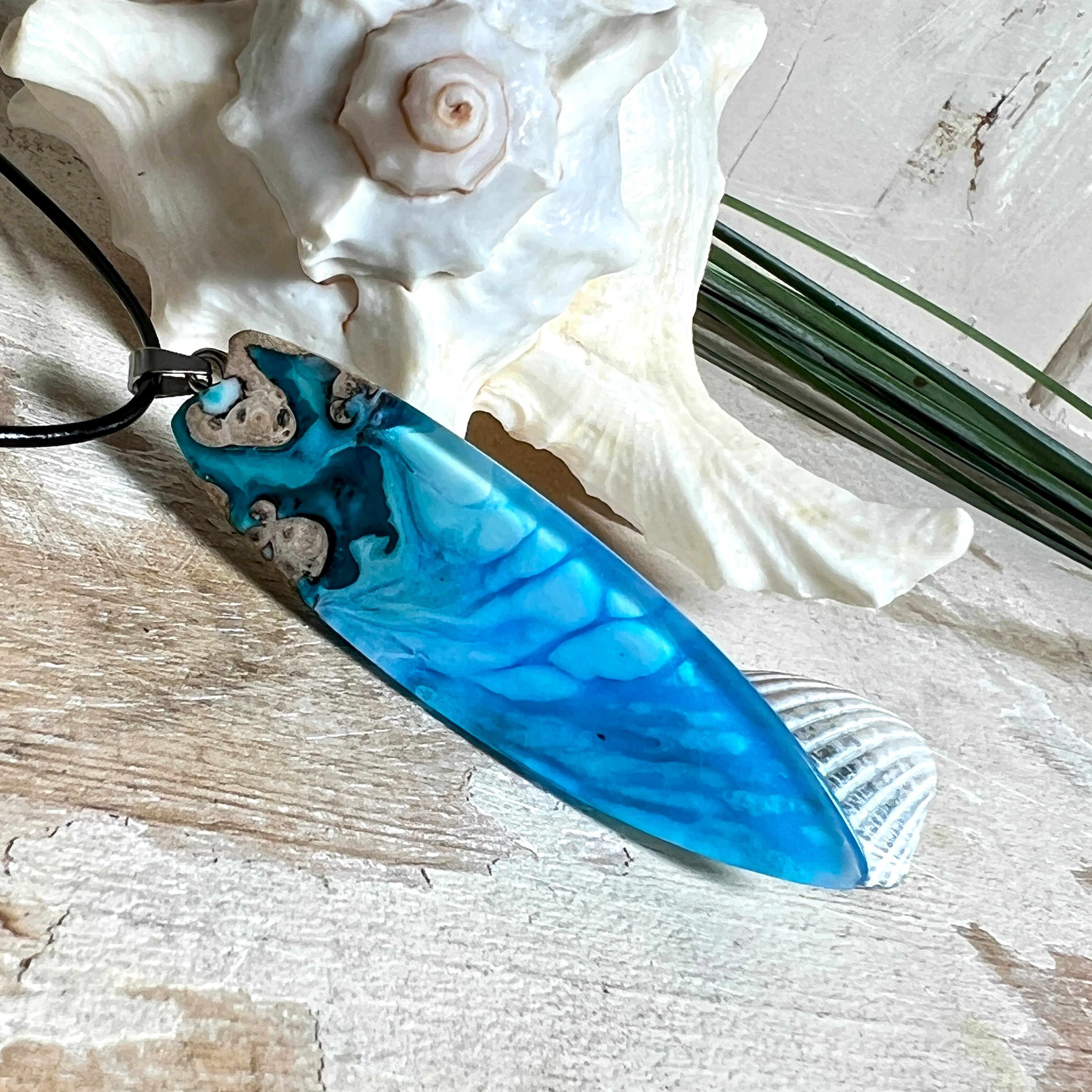 Surfbrett-Anhänger aus Maserholz und blau-weißem Epoxidharz, hochglanzpoliert, ca. 6,5 × 2 cm. An schwarzer Lederkette mit Edelstahl-Karabiner und 5-cm-Verlängerung; Edelstahl-Öse. Im Tageslicht lagunenblau, unter Schwarzlicht fluoreszierend – ideal als Party-/Festival-Schmuck. Handgefertigtes Unikat, Lieferung in edler Schmuckbox.