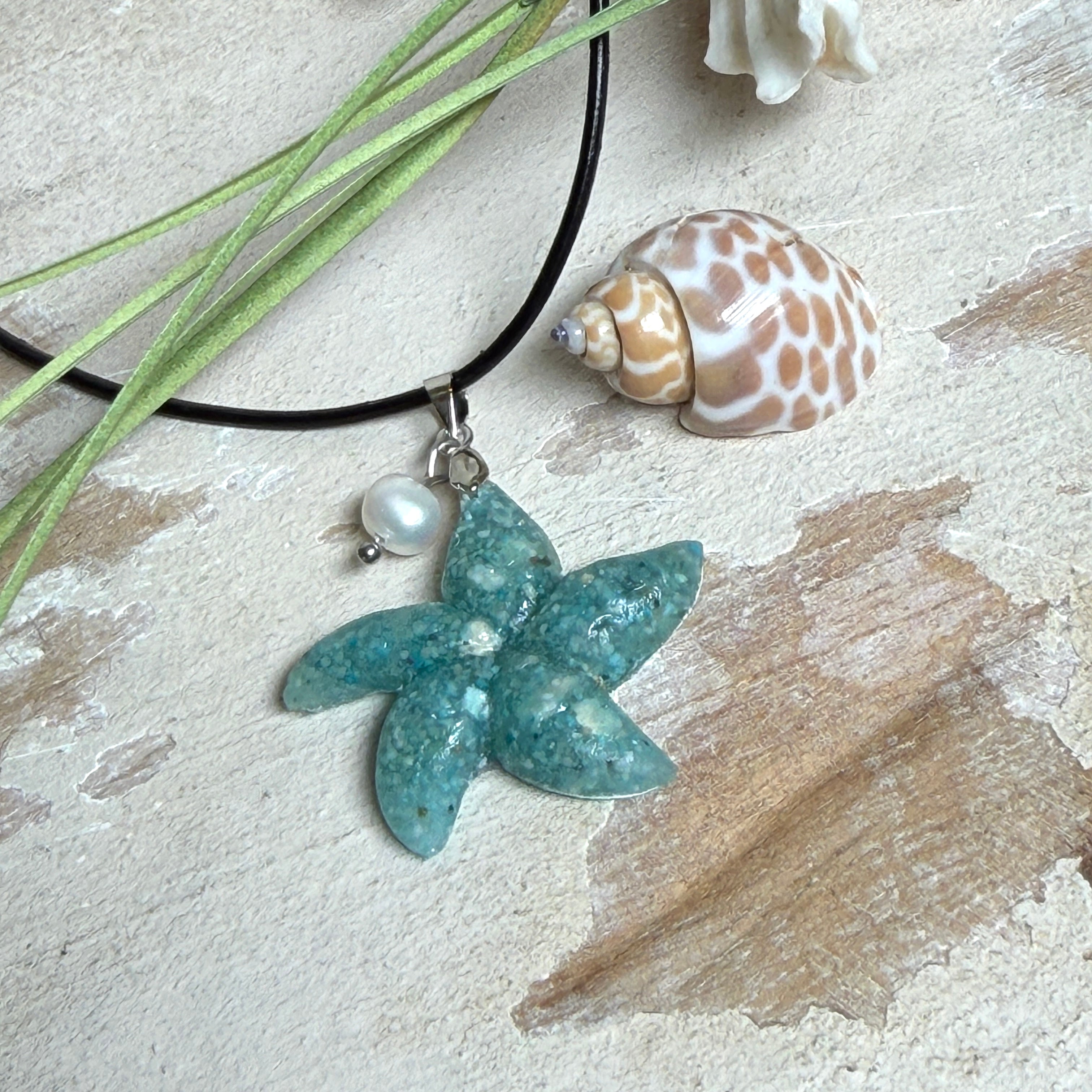 Seestern-Anhänger aus transparentem Harz mit echtem Meersand ( leicht eingefärbt), in blautürkis Strand-Look, mit kleiner Süßwasserperle als Anhänger-Detail. Edelstahlöse, schwarze Lederkette mit Edelstahl-Karabiner und Verlängerung (45 cm + 5 cm). Anhänger ca. 3,5 × 3,5 cm. Handgefertigtes Unikat; vereinzelt kleine Luftbläschen möglich, typisch für Handarbeit.