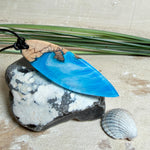 Handgefertigter Surfbrett-Anhänger aus warm gemasertem Olivenholz und Epoxidharz in Blau und Weiß, das an Meereswellen erinnert. Längliche, vorne polierte Form, ca. 6,2 × 2 cm, an verstellbarem schwarzem Lederband mit Knoten. Maritimer Unikatschmuck für Damen und Herren, dekorativ mit Stein und Muschel arrangiert.