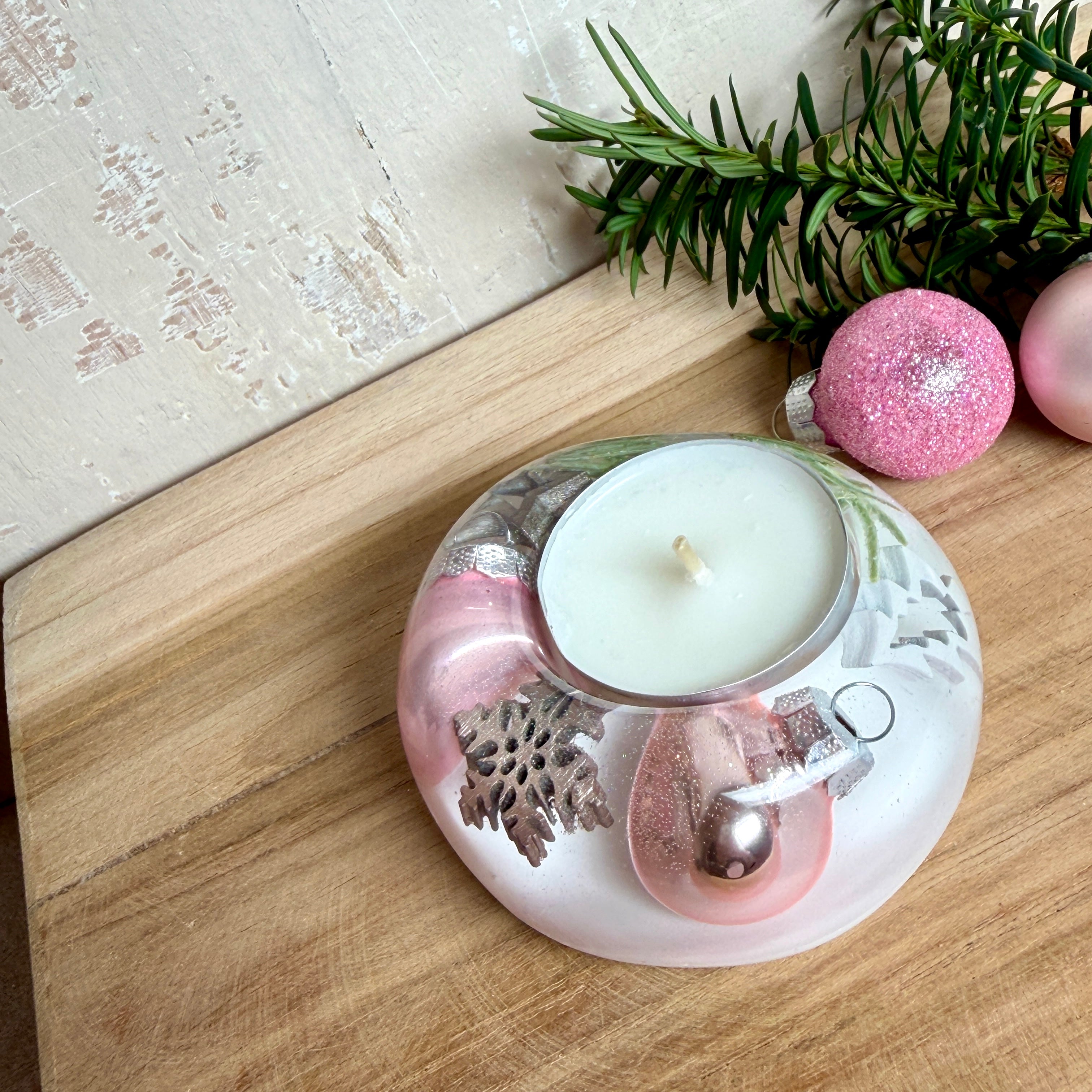 Runder Teelichthalter aus klarem Epoxidharz, Durchmesser ca. 7 cm, Höhe ca. 4 cm. Im Harz sind rosa Weihnachtskugeln, ein Stern, eine Schneeflocke, ein Tannenbaum, künstliche Tannennadeln und feiner Glitzer eingegossen. Vertiefung für ein Teelicht in der Mitte, Unterseite mit vier Korkpads als Möbelschoner. Fotografiert auf Holz mit Tannenzweig und rosa Kugeln als weihnachtliche Dekoration