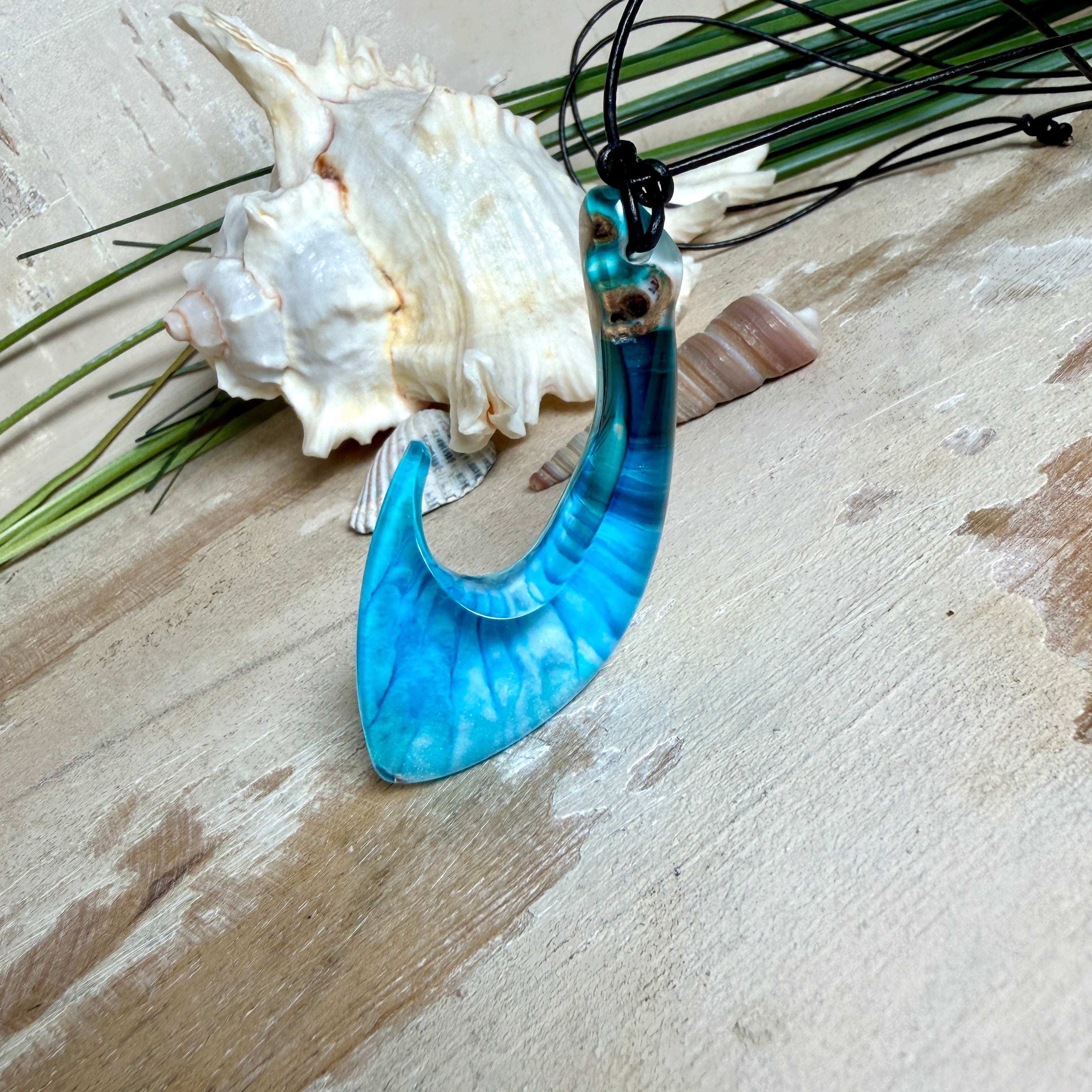 Handgefertigter Haken-Anhänger im Hei-Matau-Stil aus Maserholz und klarem Harz mit Blau-, Türkis- und Weißverlauf, hochglanzpoliert. Oben sichtbares Holz im Harzbereich, getragen an verstellbarem schwarzem Lederband mit Schiebeknoten und Zierperlen. Anhänger ca. 7 × 3 cm, fotografiert mit Muscheln im Hintergrund.