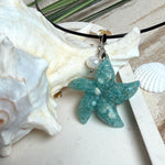 Seestern-Anhänger aus transparentem Harz mit echtem Meersand ( leicht eingefärbt), in blautürkis Strand-Look, mit kleiner Süßwasserperle als Anhänger-Detail. Edelstahlöse, schwarze Lederkette mit Edelstahl-Karabiner und Verlängerung (45 cm + 5 cm). Anhänger ca. 3,5 × 3,5 cm. Handgefertigtes Unikat; vereinzelt kleine Luftbläschen möglich, typisch für Handarbeit.