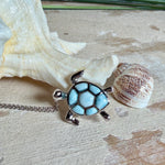 Feine Kette aus rhodiniertem 925 Silber mit Wasserschildkröten-Anhänger. Der ca. 25 x 18 mm große Anhänger zeigt eine Schildkröte, deren Panzer aus mehreren hellblauen Larimar-Inlays besteht. Seltener, kostbarer Larimar mit typischen wolkigen Strukturen, an einer ca. 45 cm langen, 1 mm starken, auf Platin rhodinierten Silberkette. Fotografiert in schwarzer Schmuckbox und vor maritimer Dekoration mit Muscheln.