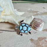 Feine Kette aus rhodiniertem 925 Silber mit Wasserschildkröten-Anhänger. Der ca. 25 x 18 mm große Anhänger zeigt eine Schildkröte, deren Panzer aus mehreren hellblauen Larimar-Inlays besteht. Seltener, kostbarer Larimar mit typischen wolkigen Strukturen, an einer ca. 45 cm langen, 1 mm starken, auf Platin rhodinierten Silberkette. Fotografiert in schwarzer Schmuckbox und vor maritimer Dekoration mit Muscheln.