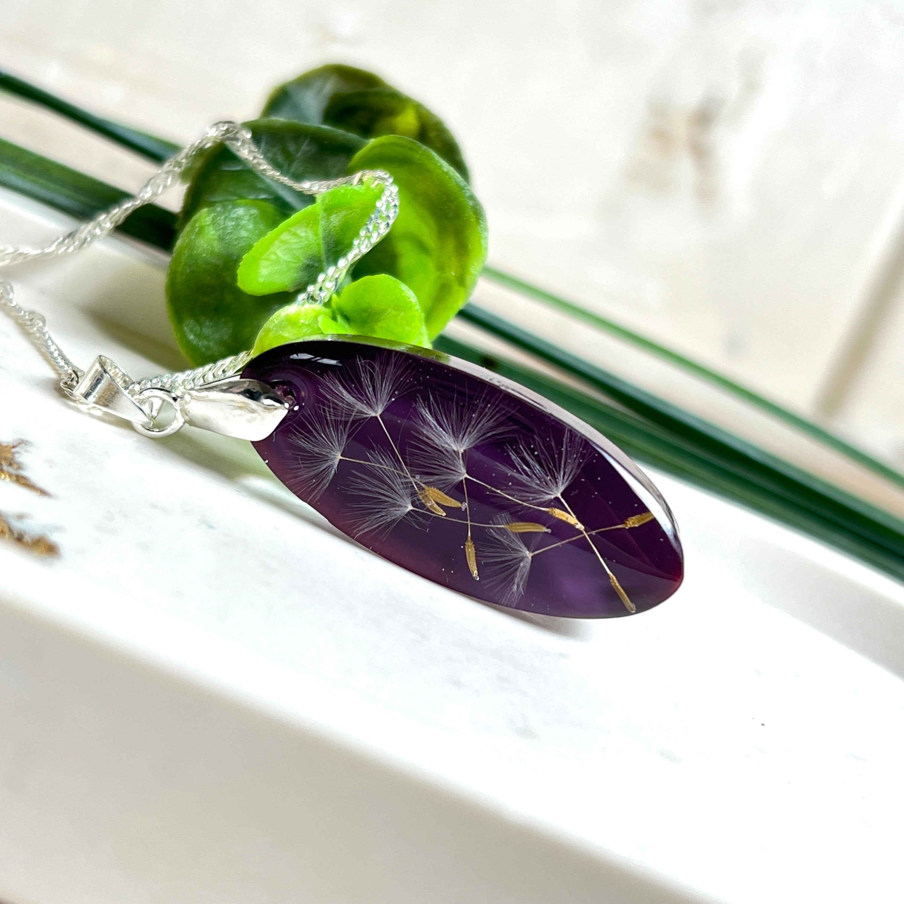 Ovaler Anhänger mit echter Pusteblume in Harz, Hintergrund Brombeerfarben. Größe 4 × 1,8 cm, versilberte Kette 50 cm mit Karabinerverschluss. Handgefertigtes Unikat.