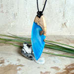 Handgefertigter Surfbrett-Anhänger aus warm gemasertem Olivenholz und Epoxidharz in Blau und Weiß, das an Meereswellen erinnert. Längliche, vorne polierte Form, ca. 6,2 × 2 cm, an verstellbarem schwarzem Lederband mit Knoten. Maritimer Unikatschmuck für Damen und Herren, dekorativ mit Stein und Muschel arrangiert.