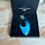 Handgefertigter Haken-Anhänger im Hei-Matau-Stil aus Maserholz und klarem Harz mit Blau-, Türkis- und Weißverlauf, hochglanzpoliert. Oben sichtbares Holz im Harzbereich, getragen an verstellbarem schwarzem Lederband mit Schiebeknoten und Zierperlen. Anhänger ca. 7 × 3 cm, fotografiert mit Muscheln im Hintergrund.