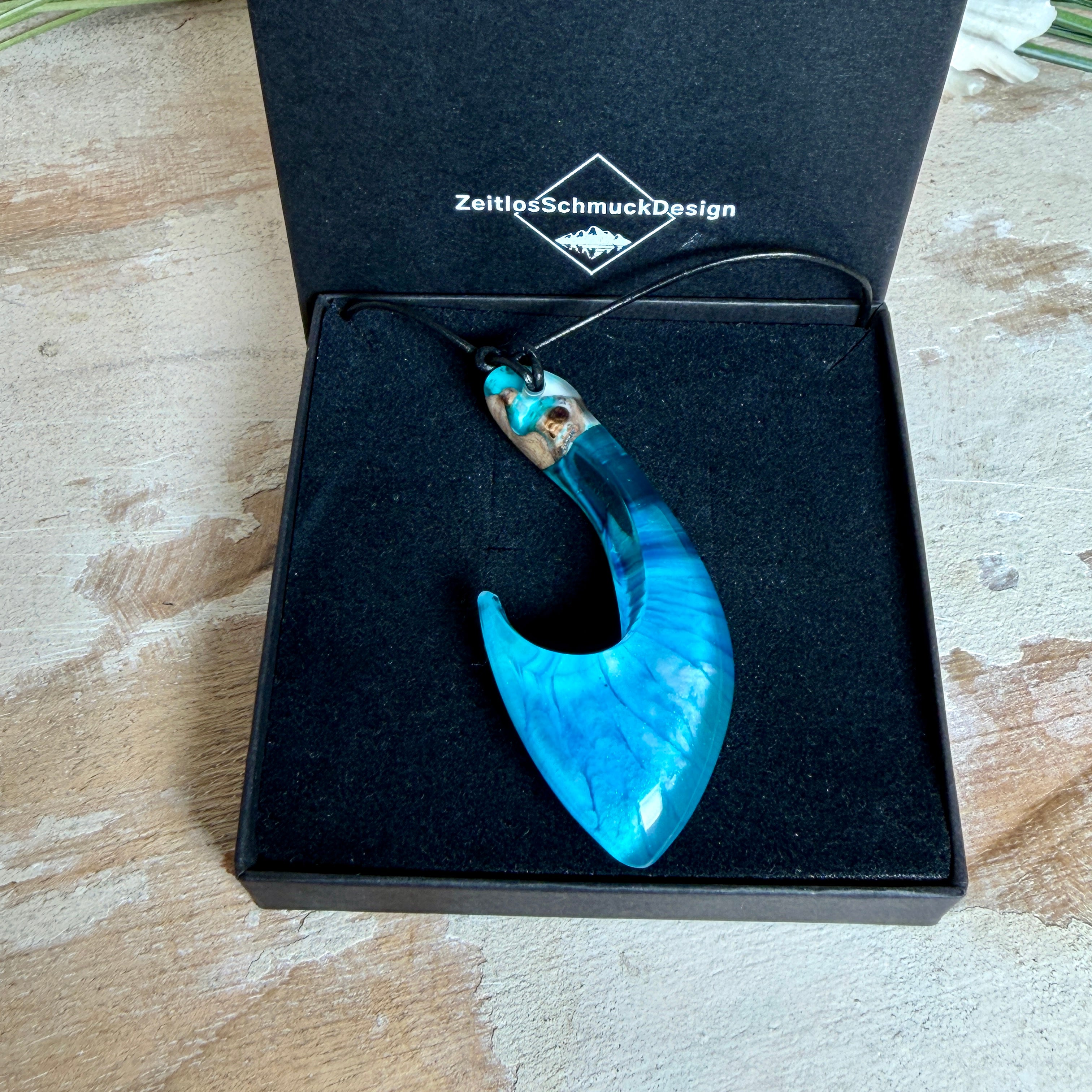 Handgefertigter Haken-Anhänger im Hei-Matau-Stil aus Maserholz und klarem Harz mit Blau-, Türkis- und Weißverlauf, hochglanzpoliert. Oben sichtbares Holz im Harzbereich, getragen an verstellbarem schwarzem Lederband mit Schiebeknoten und Zierperlen. Anhänger ca. 7 × 3 cm, fotografiert mit Muscheln im Hintergrund.