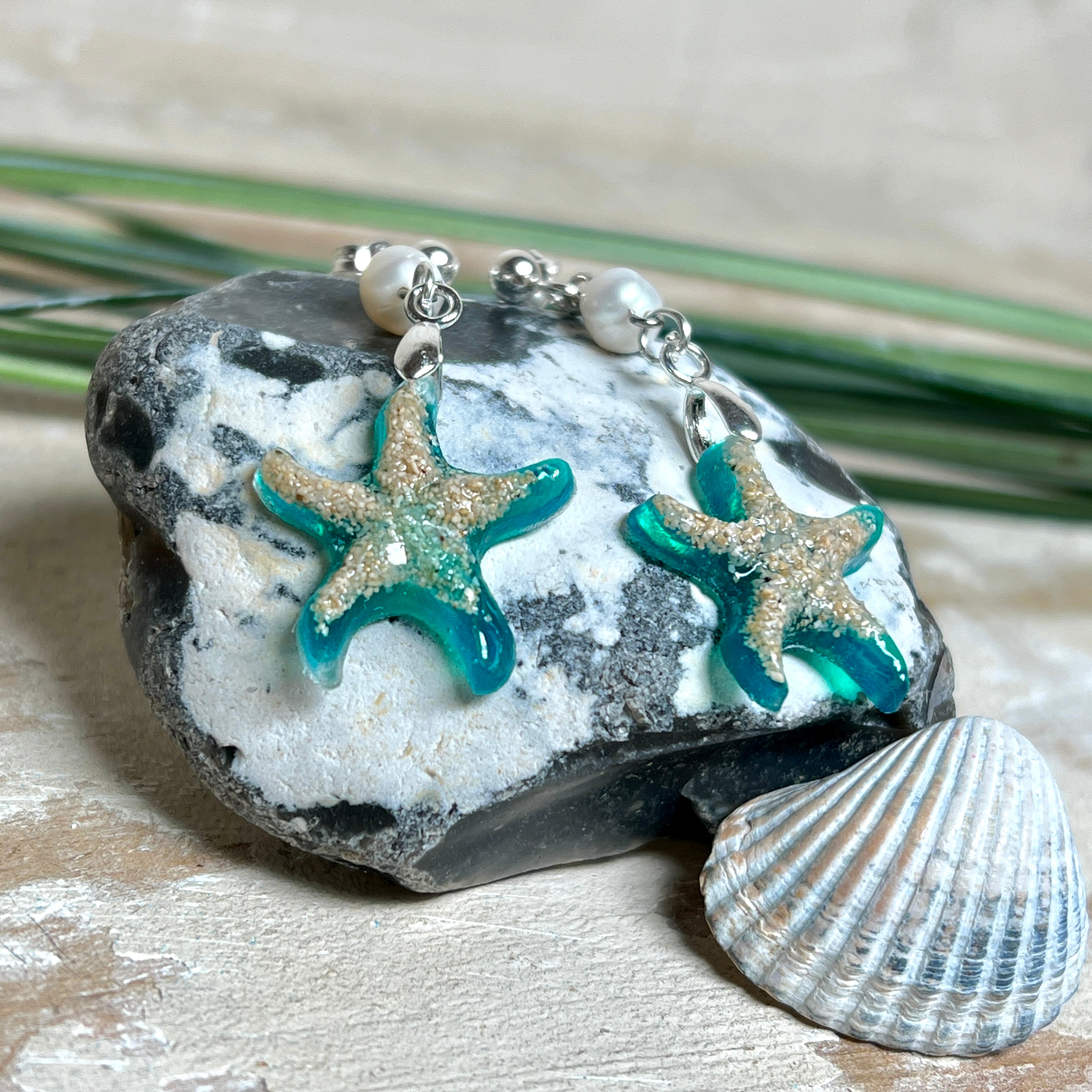 Handgefertigte Seestern-Ohrringe aus Epoxidharz und Sand mit Süßwasserperle, an 925 Sterling Silber Ohrsteckern; versilberte Anhängerschlaufe. Je Seestern ca. 2,6 × 2,5 cm. Leichte, maritime Dangle-Ohrringe im Strand-Look – ideal als Geschenk für Meeresliebhaberinnen.