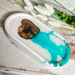 Handgefertigtes Schmucktablett aus Jesmonite mit eingebettetem Olivenholz und türkisfarbenem Resin. Die glänzende Oberfläche erinnert an Wasserstrukturen, während das Naturholz Tiefe und Wärme verleiht. Die Rückseite ist mit Korkpunkten versehen und schützt empfindliche Oberflächen. Perfekt als Ablage für Schmuck, Brille, Schlüssel oder Deko jedes Stück ein Unikat.