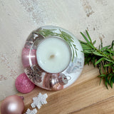 Runder Teelichthalter aus klarem Epoxidharz, Durchmesser ca. 7 cm, Höhe ca. 4 cm. Im Harz sind rosa Weihnachtskugeln, ein Stern, eine Schneeflocke, ein Tannenbaum, künstliche Tannennadeln und feiner Glitzer eingegossen. Vertiefung für ein Teelicht in der Mitte, Unterseite mit vier Korkpads als Möbelschoner. Fotografiert auf Holz mit Tannenzweig und rosa Kugeln als weihnachtliche Dekoration