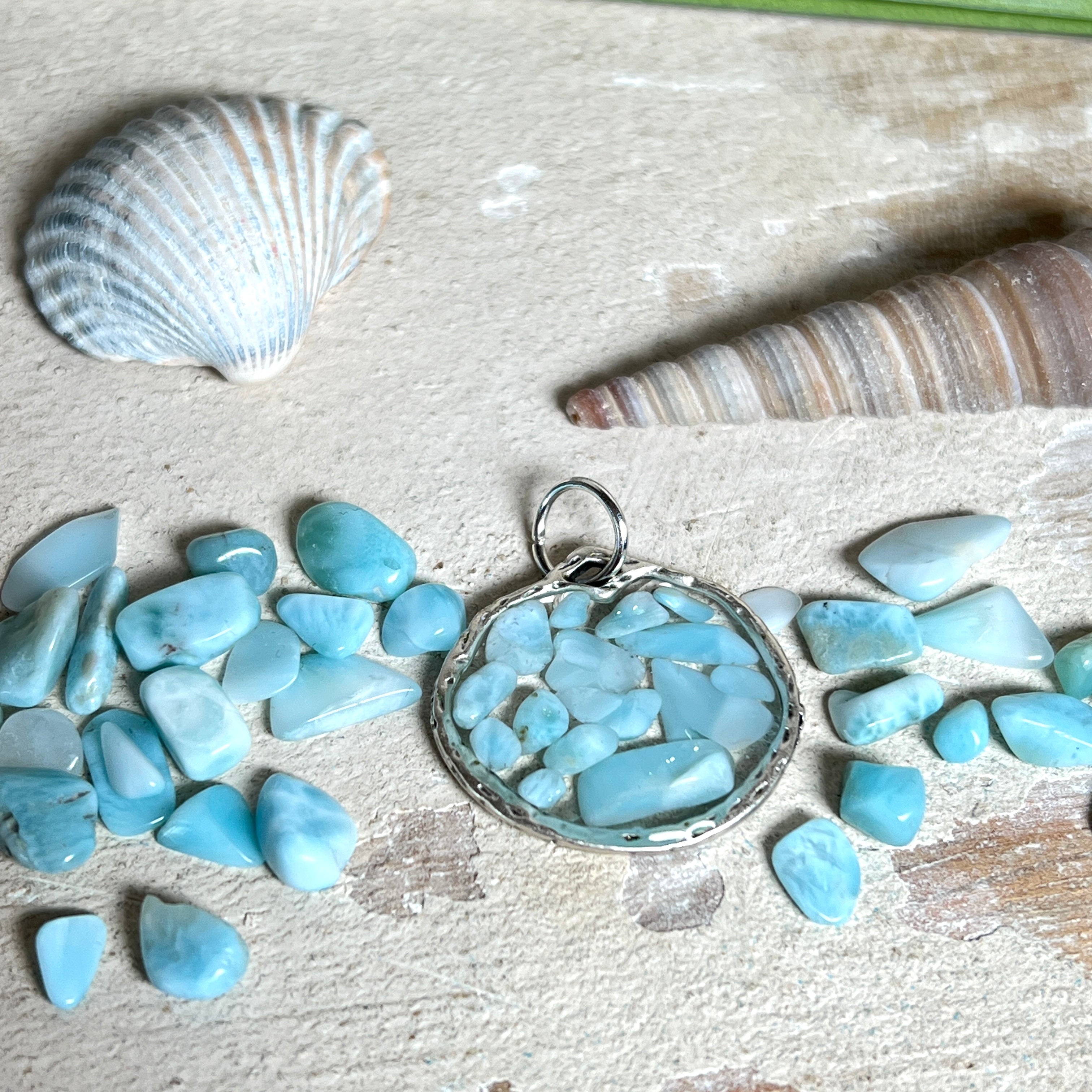 Runder Anhänger „Atlantis Dream“ aus antik-silberfarbener Zinklegierung, gefüllt mit kleinen Larimar-Edelsteinchen und klarem Resin. Die Larimarsteine zeigen sanfte Blau- und Weißtöne, die an das Meer erinnern. Durchmesser ca. 22 mm, handgefertigtes Unikat, fotografiert auf maritimem Untergrund mit kleinen Larimarsteinen und Muscheln zur Größenorientierung.