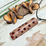 Handgefertigter Halsanhänger aus echtem Banksiazapfen, veredelt mit rotem Harz in den natürlichen Kammern; ca. 5,5 × 1,6 cm. Mit Anhängerschlaufe aus Edelstahl an schwarzer Lederkette (45 cm + 5 cm Verlängerung) mit Edelstahl-Karabiner. Detailaufnahme bzw. Tragebild vor neutralem, natürlichem Hintergrund.