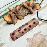 Handgefertigter Halsanhänger aus echtem Banksiazapfen, veredelt mit rotem Harz in den natürlichen Kammern; ca. 5,5 × 1,6 cm. Mit Anhängerschlaufe aus Edelstahl an schwarzer Lederkette (45 cm + 5 cm Verlängerung) mit Edelstahl-Karabiner. Detailaufnahme bzw. Tragebild vor neutralem, natürlichem Hintergrund.