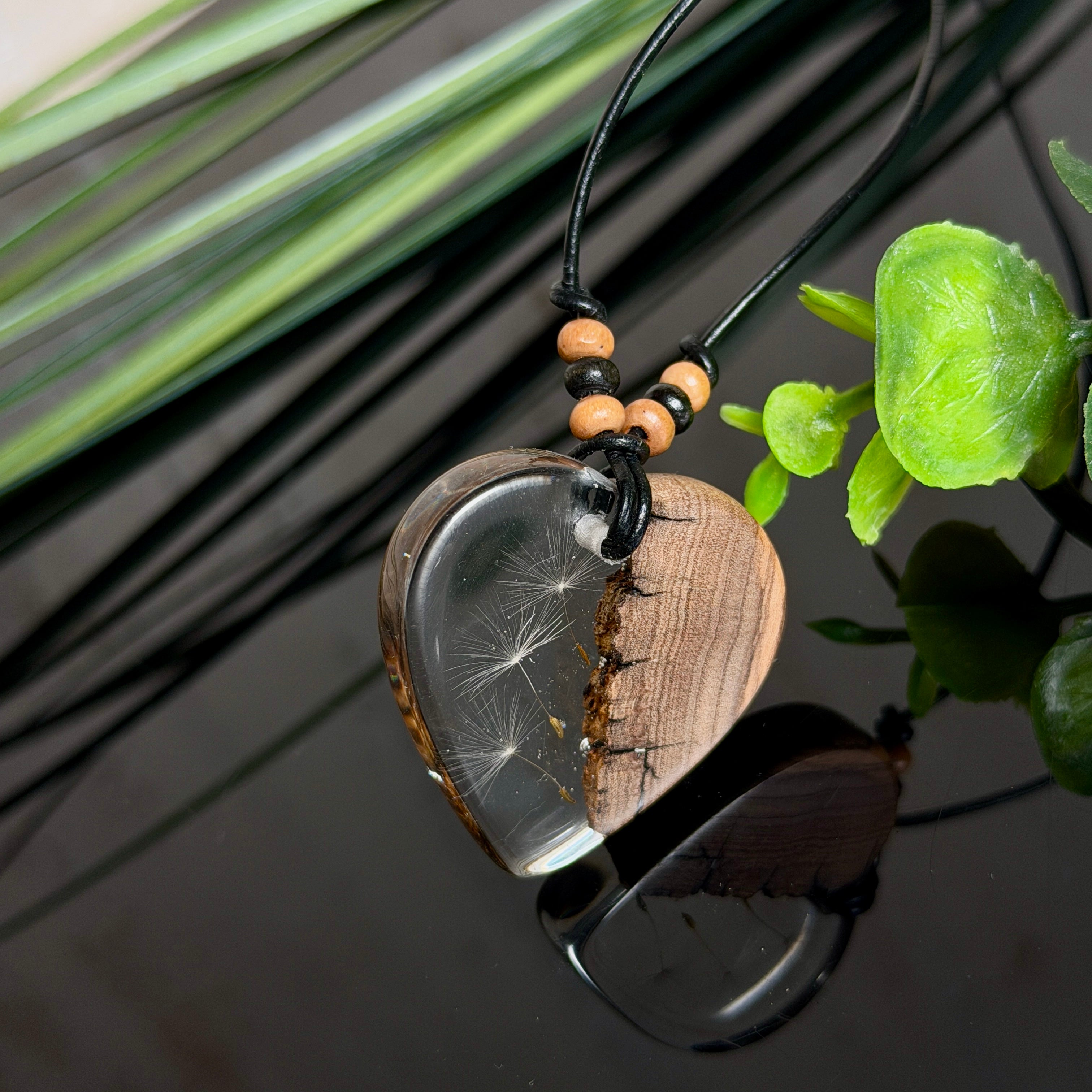 Handgefertigter Herzanhänger aus warm gemasertem Olivenholz und kristallklarem Epoxidharz mit schwebenden Pusteblumensamen. Schwarzes, verstellbares Lederband mit Schiebeknoten und naturfarbenen Holzperlen, Anhänger ca. 3,5 × 3,3 cm. Naturnaher Unikatschmuck von ZeitlosSchmuckDesign.