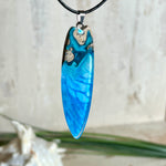 Surfbrett-Anhänger aus Maserholz und blau-weißem Epoxidharz, hochglanzpoliert, ca. 6,5 × 2 cm. An schwarzer Lederkette mit Edelstahl-Karabiner und 5-cm-Verlängerung; Edelstahl-Öse. Im Tageslicht lagunenblau, unter Schwarzlicht fluoreszierend – ideal als Party-/Festival-Schmuck. Handgefertigtes Unikat, Lieferung in edler Schmuckbox.