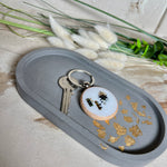 Handgefertigtes ovales Schmucktablett aus grauen  Jesmonite mit schimmernden Blattgold-Details. Die Unterseite ist mit vier Korkpunkten versehen und schützt empfindliche Oberflächen. Ideal zur stilvollen Aufbewahrung von Schmuck, Schlüsseln, Brille oder kleinen Dekoartikeln. Jedes Tablett ist ein Unikat mit individueller Maserung und goldenen Akzenten – Lieferung ohne Deko. Die 