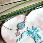 Verstellbares Edelstahlarmband mit Zirkoniasteinchen und Schiebeverschluss und rundem Anhänger (ca. 2,5 × 1,7 cm) mit echten Larimar-Edelstein-Splittern in Harz. Zusätzlich ein Charm „Blume des Lebens“ aus 925 Silber. Handgefertigtes Unikat in Türkis- und Meeresblautönen – Schmuckstück in Nahaufnahme bzw. als Tragebild am Handgelenk.