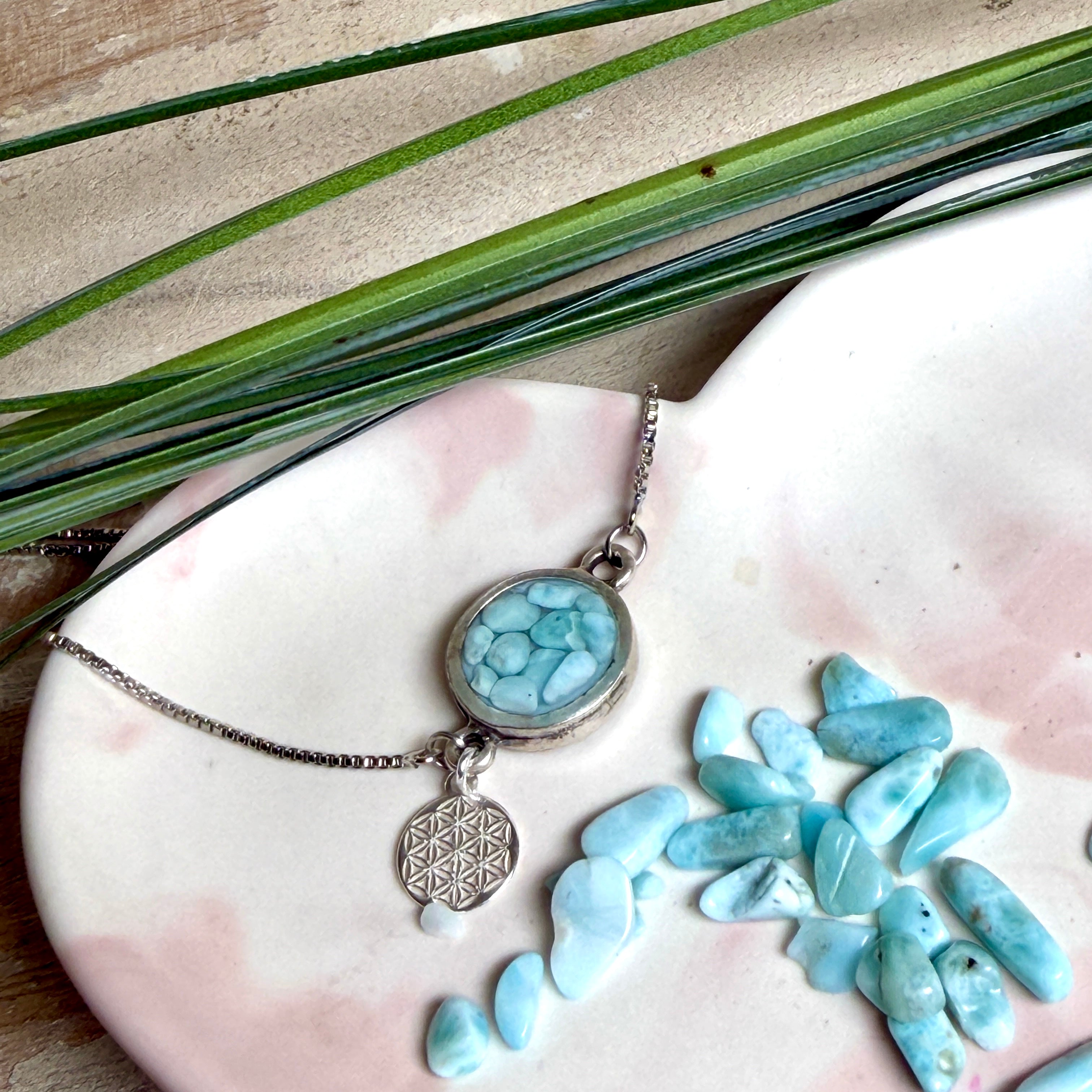 Verstellbares Edelstahlarmband mit Zirkoniasteinchen und Schiebeverschluss und rundem Anhänger (ca. 2,5 × 1,7 cm) mit echten Larimar-Edelstein-Splittern in Harz. Zusätzlich ein Charm „Blume des Lebens“ aus 925 Silber. Handgefertigtes Unikat in Türkis- und Meeresblautönen – Schmuckstück in Nahaufnahme bzw. als Tragebild am Handgelenk.