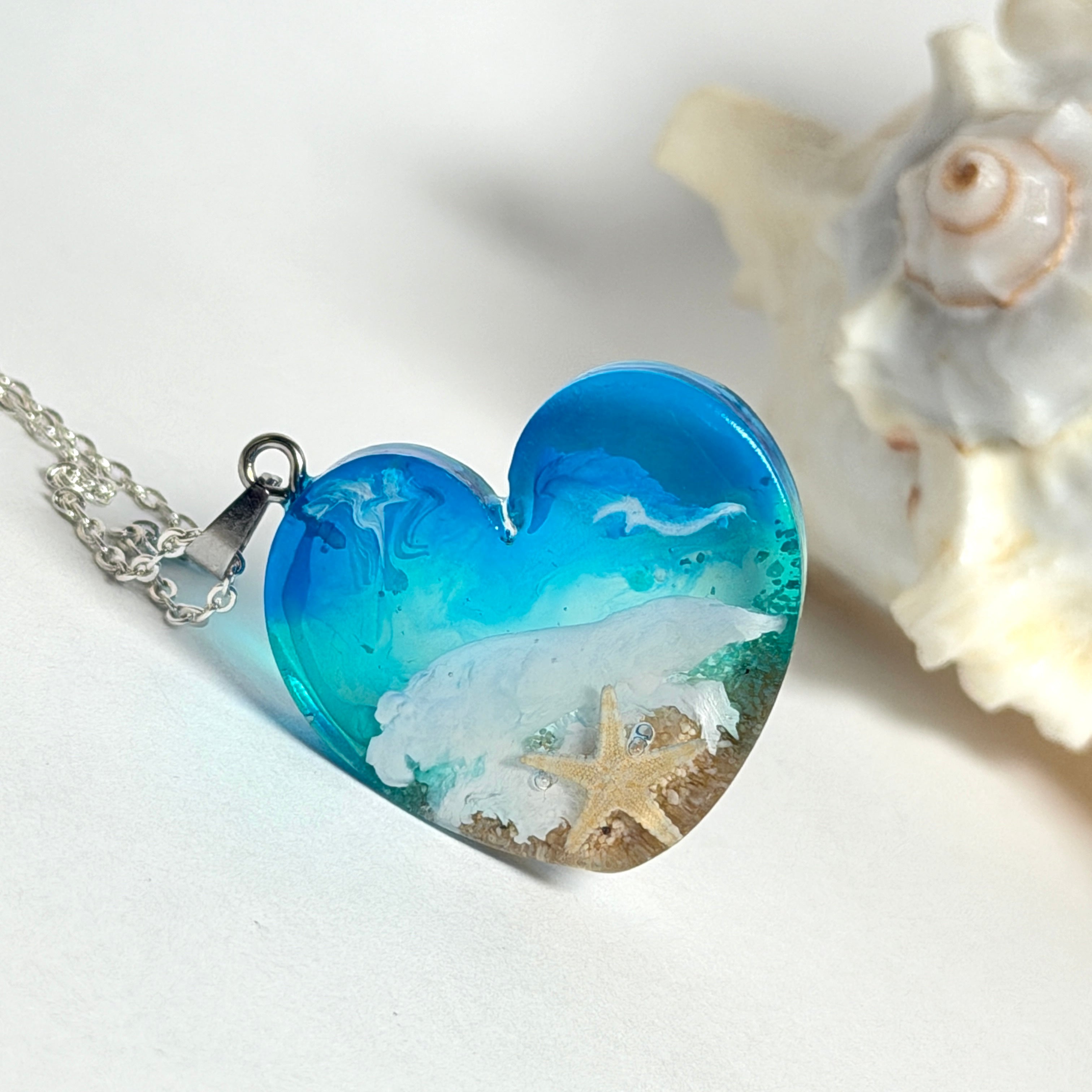 Ein herzförmiger Anhänger mit einer ganzen Ozeanwelt darin: Welle, Sand und Seestern in leuchtendem Blau und Türkis – handgegossen, einzigartig, unvergesslich. Mit versilberter Kette (46 cm) und Edelstahl-Karabiner.