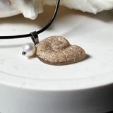 Handgefertigter Ammonit-Anhänger aus echtem Meersand & Harz mit filigraner Edelstahlöse und einer edlen Süßwasserperle. Maritimer Schmuck, inspiriert von der Kraft und Schönheit des Ozeans. Jedes Stück ein Unikat. Präsentiert auf einer Muschel, mit Lederkette.