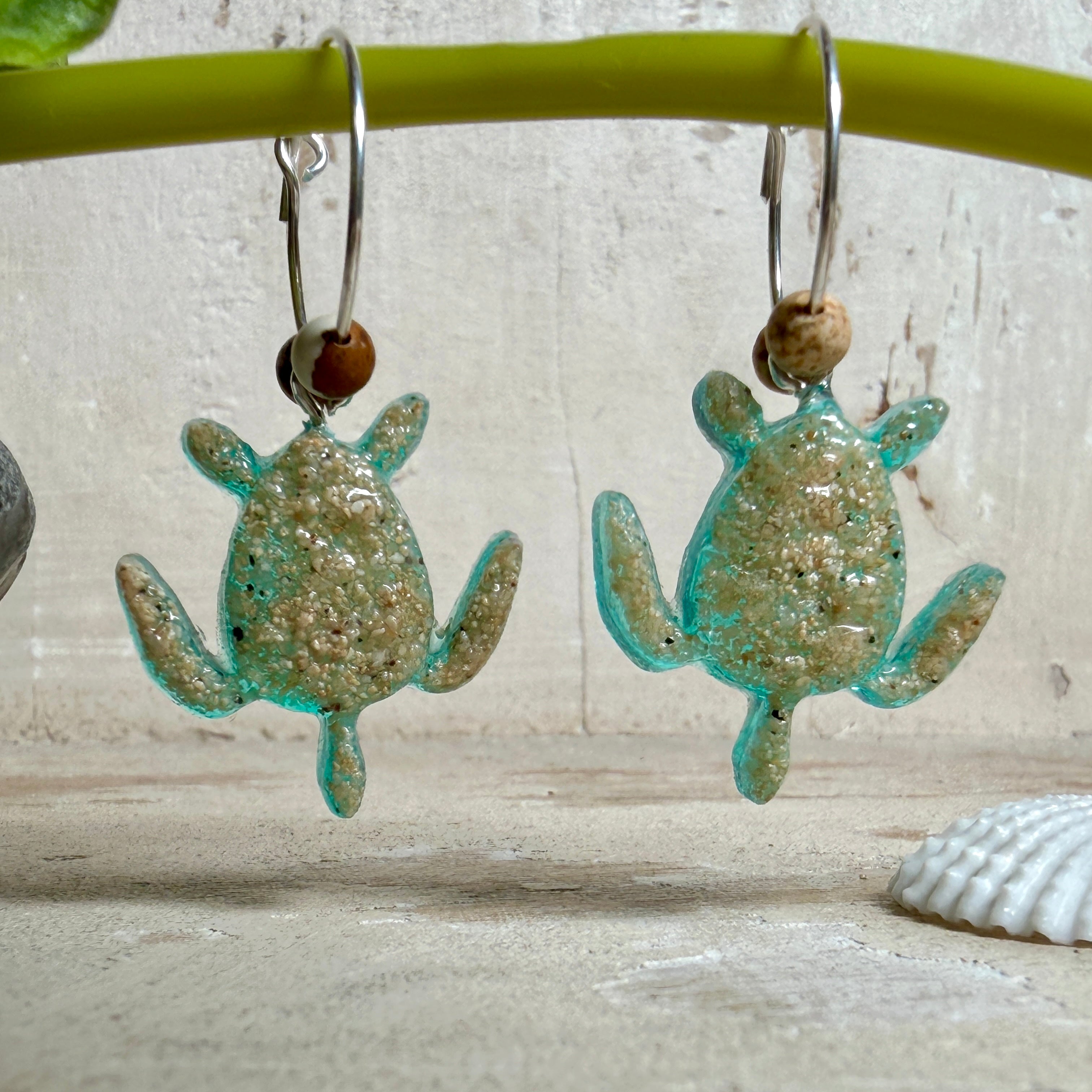 Türkisfarbene Schildkröten-Creolen an feinen 16-mm-Ringen aus 925 Silber, jeweils mit echtem Strandsand im Harz eingebettet und mit kleinen Jaspis-Edelsteinperlen verziert; handgefertigtes Unikat mit sommerlich-maritimem Look, fotografiert als Freisteller, Nahaufnahme oder auf dekorativem Untergrund.