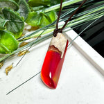 Handgefertigter Hei Toki Anhänger aus Goldfield-Maserholz und rubinrotem Labor-Opal, eingebettet in glänzendes Epoxidharz. Je nach Lichteinfall schimmert das Harz in warmen Farben mit Regenbogeneffekt. Der Anhänger ist ca. 4,3 × 1,5 cm groß und wird an einem verstellbaren Lederband mit Holzperlen getragen. Symbolischer Unikatschmuck – ideal als Geschenk für besondere Menschen.