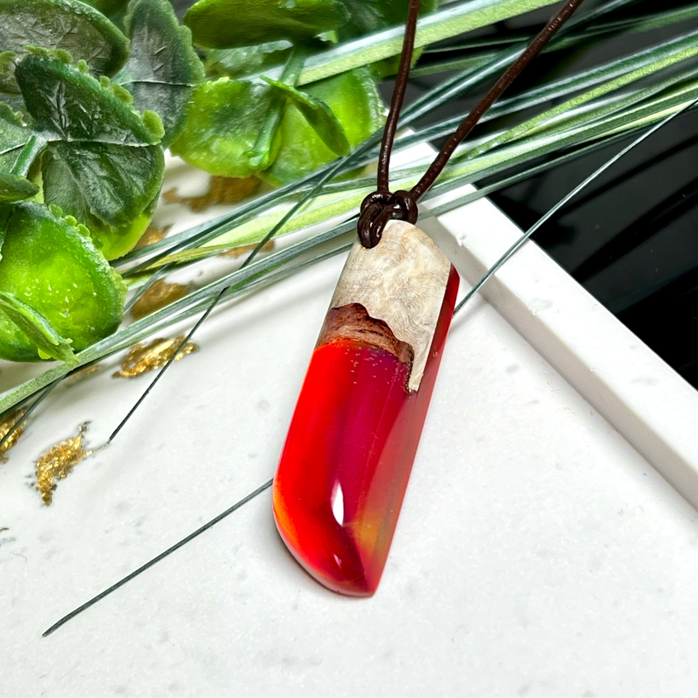 Handgefertigter Hei Toki Anhänger aus Goldfield-Maserholz und rubinrotem Labor-Opal, eingebettet in glänzendes Epoxidharz. Je nach Lichteinfall schimmert das Harz in warmen Farben mit Regenbogeneffekt. Der Anhänger ist ca. 4,3 × 1,5 cm groß und wird an einem verstellbaren Lederband mit Holzperlen getragen. Symbolischer Unikatschmuck – ideal als Geschenk für besondere Menschen.
