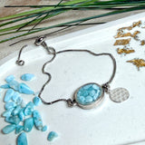 Verstellbares Edelstahlarmband mit Zirkoniasteinchen und Schiebeverschluss und rundem Anhänger (ca. 2,5 × 1,7 cm) mit echten Larimar-Edelstein-Splittern in Harz. Zusätzlich ein Charm „Blume des Lebens“ aus 925 Silber. Handgefertigtes Unikat in Türkis- und Meeresblautönen – Schmuckstück in Nahaufnahme bzw. als Tragebild am Handgelenk.