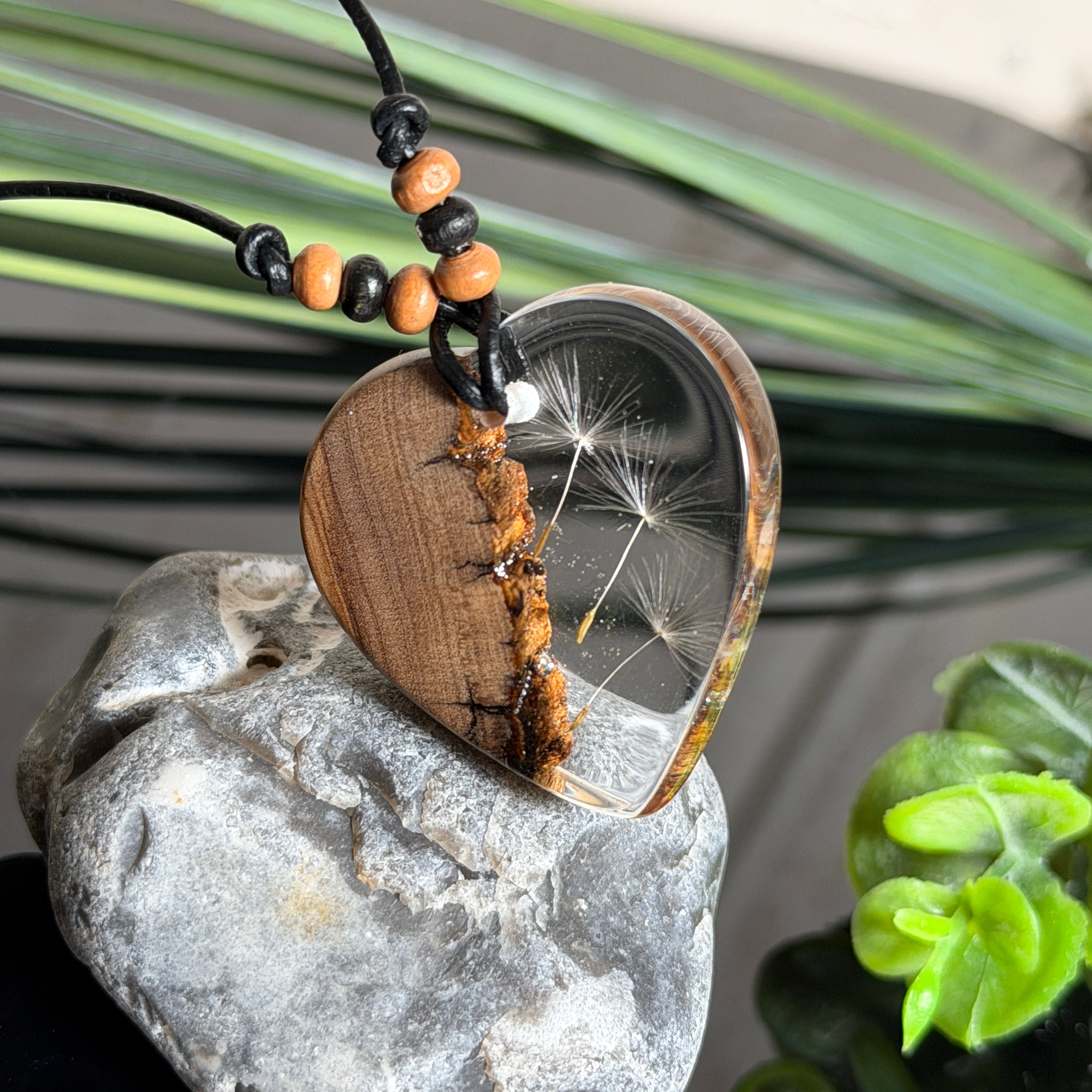 Handgefertigter Herzanhänger aus warm gemasertem Olivenholz und kristallklarem Epoxidharz mit schwebenden Pusteblumensamen. Schwarzes, verstellbares Lederband mit Schiebeknoten und naturfarbenen Holzperlen, Anhänger ca. 3,5 × 3,3 cm. Naturnaher Unikatschmuck von ZeitlosSchmuckDesign.