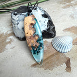 Handgefertigter, länglicher Surfbrett-Anhänger aus naturbelassenem Maserholz und transparentem Harz in Lagunen-Türkis, an schwarzem Lederband mit Knoten – maritimer Unikatschmuck für Damen und Herren, dekorativ mit Stein und Muschel inszeniert.
