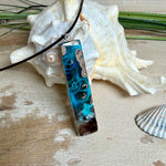 Rechteckiger Stabanhänger aus Maserholz und glasklarem Epoxidharz in tiefen Blau- und Türkistönen mit organischer Lagunen/Riff-Optik. Hochglanzpoliert, ca. 5,3 × 1,2 cm. Der Anhänger hängt an einer schwarzen Lederkette mit Edelstahl-Karabiner und 5-cm-Verlängerung (45 + 5 cm); die Anhängerschlaufe ist ebenfalls aus Edelstahl. Handgefertigtes Unikat, Lieferung in edler Schmuckbox – maritimer Look.