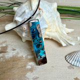 Rechteckiger Stabanhänger aus Maserholz und glasklarem Epoxidharz in tiefen Blau- und Türkistönen mit organischer Lagunen/Riff-Optik. Hochglanzpoliert, ca. 5,3 × 1,2 cm. Der Anhänger hängt an einer schwarzen Lederkette mit Edelstahl-Karabiner und 5-cm-Verlängerung (45 + 5 cm); die Anhängerschlaufe ist ebenfalls aus Edelstahl. Handgefertigtes Unikat, Lieferung in edler Schmuckbox – maritimer Look.