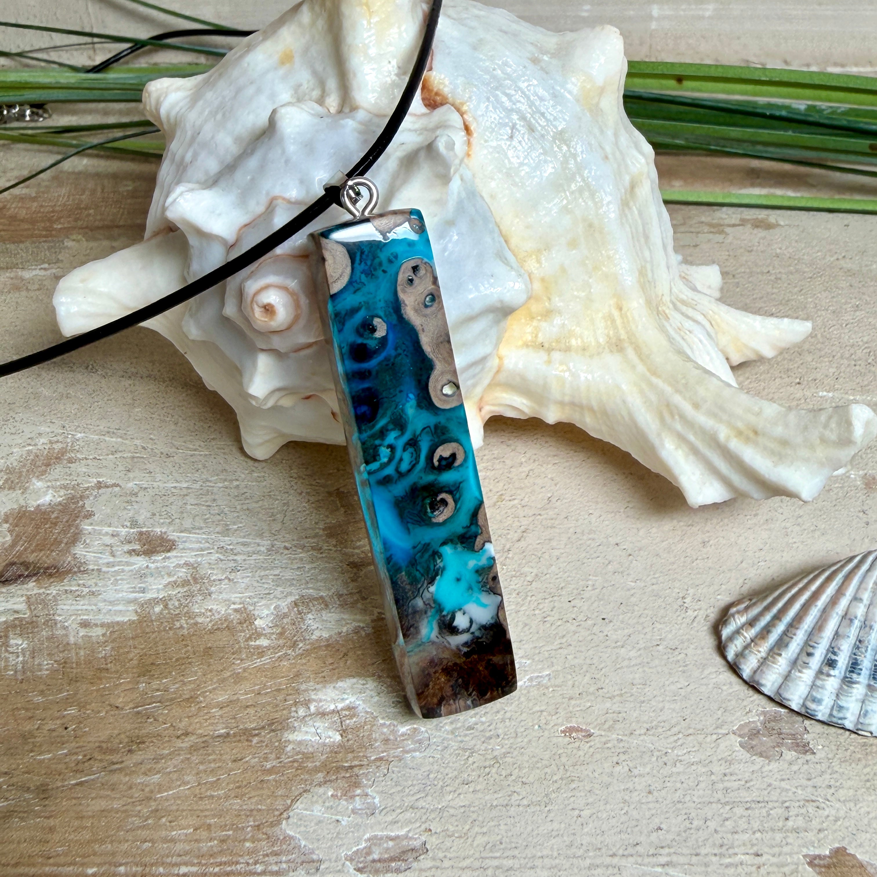 Rechteckiger Stabanhänger aus Maserholz und glasklarem Epoxidharz in tiefen Blau- und Türkistönen mit organischer Lagunen/Riff-Optik. Hochglanzpoliert, ca. 5,3 × 1,2 cm. Der Anhänger hängt an einer schwarzen Lederkette mit Edelstahl-Karabiner und 5-cm-Verlängerung (45 + 5 cm); die Anhängerschlaufe ist ebenfalls aus Edelstahl. Handgefertigtes Unikat, Lieferung in edler Schmuckbox – maritimer Look.