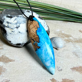 Handgefertigter Surfbrett-Anhänger aus warm gemasertem Olivenholz und Epoxidharz in Blau und Weiß, das an Meereswellen erinnert. Längliche, vorne polierte Form, ca. 6,2 × 2 cm, an verstellbarem schwarzem Lederband mit Knoten. Maritimer Unikatschmuck für Damen und Herren, dekorativ mit Stein und Muschel arrangiert.