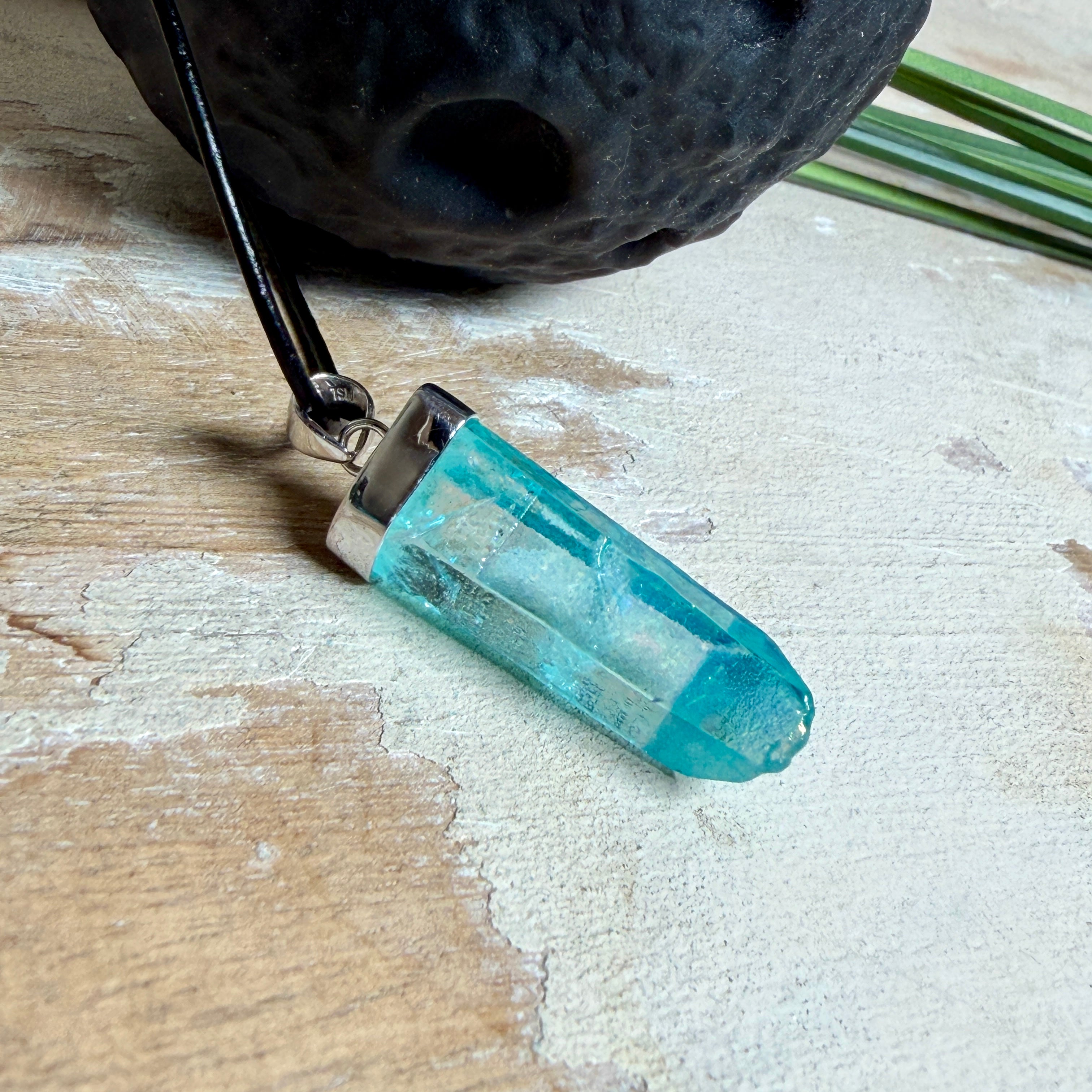Nahaufnahme einer handgefertigten Halskette mit Aqua Aura Rohkristall: naturbelassener, sechskantiger Bergkristall mit goldbedampfter Aqua-Beschichtung in Türkisblau, gefasst in 925er Silber und getragen an einer schwarzen Lederkette mit Edelstahl-Karabiner und Verlängerungskettchen.