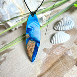 Handgefertigter Surfbrett-Anhänger „Ocean Glide“ aus Maserholz und Epoxidharz in verschiedenen Blau-, und Weißtönen. Der ca. 6,5 × 2 cm große Anhänger wird an einem verstellbaren Lederband (26–46 cm) mit Holzperlen geliefert. Ein maritimes Unikat mit natürlicher Holzmaserung, das die Schönheit des Meeres einfängt.