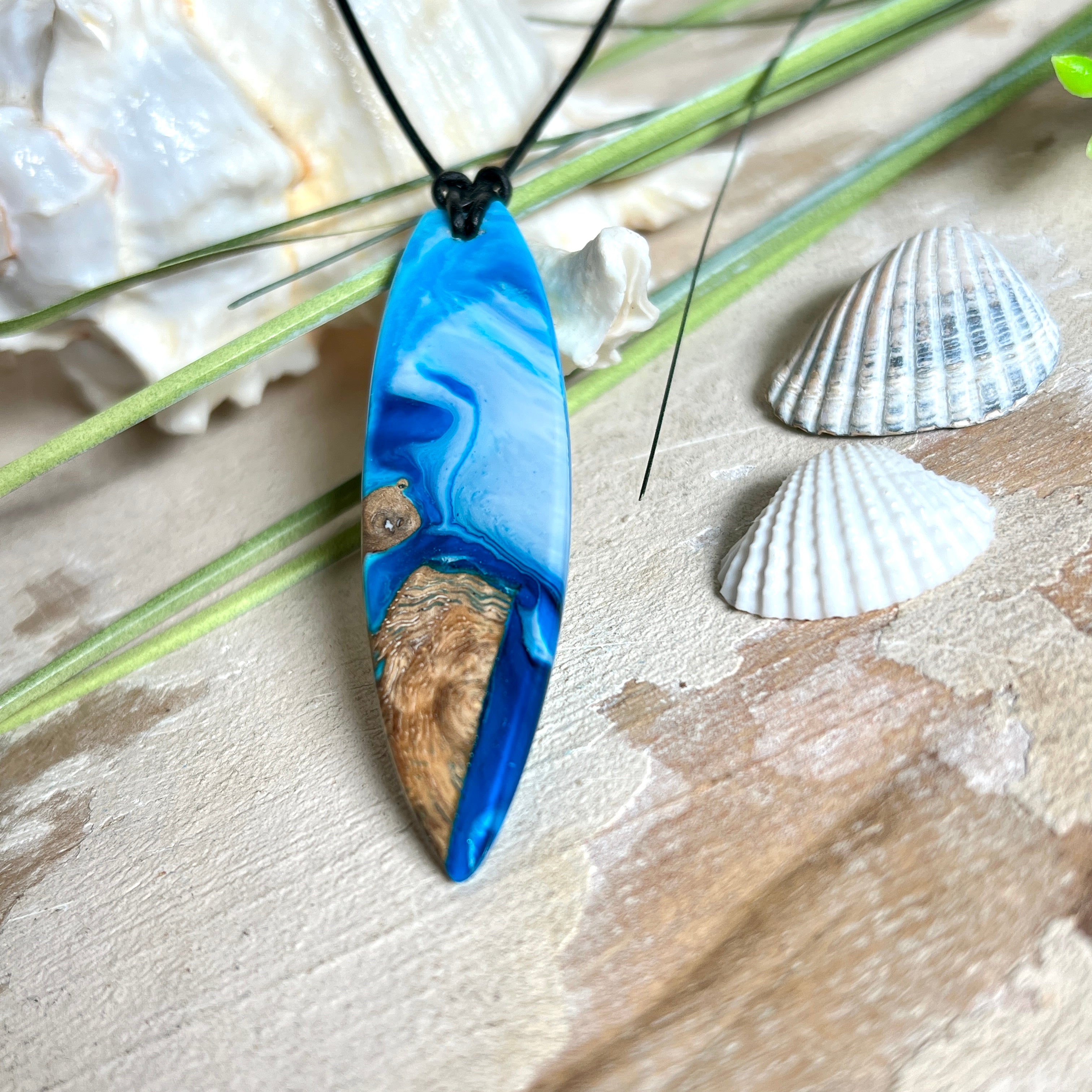 Handgefertigter Surfbrett-Anhänger „Ocean Glide“ aus Maserholz und Epoxidharz in verschiedenen Blau-, und Weißtönen. Der ca. 6,5 × 2 cm große Anhänger wird an einem verstellbaren Lederband (26–46 cm) mit Holzperlen geliefert. Ein maritimes Unikat mit natürlicher Holzmaserung, das die Schönheit des Meeres einfängt.