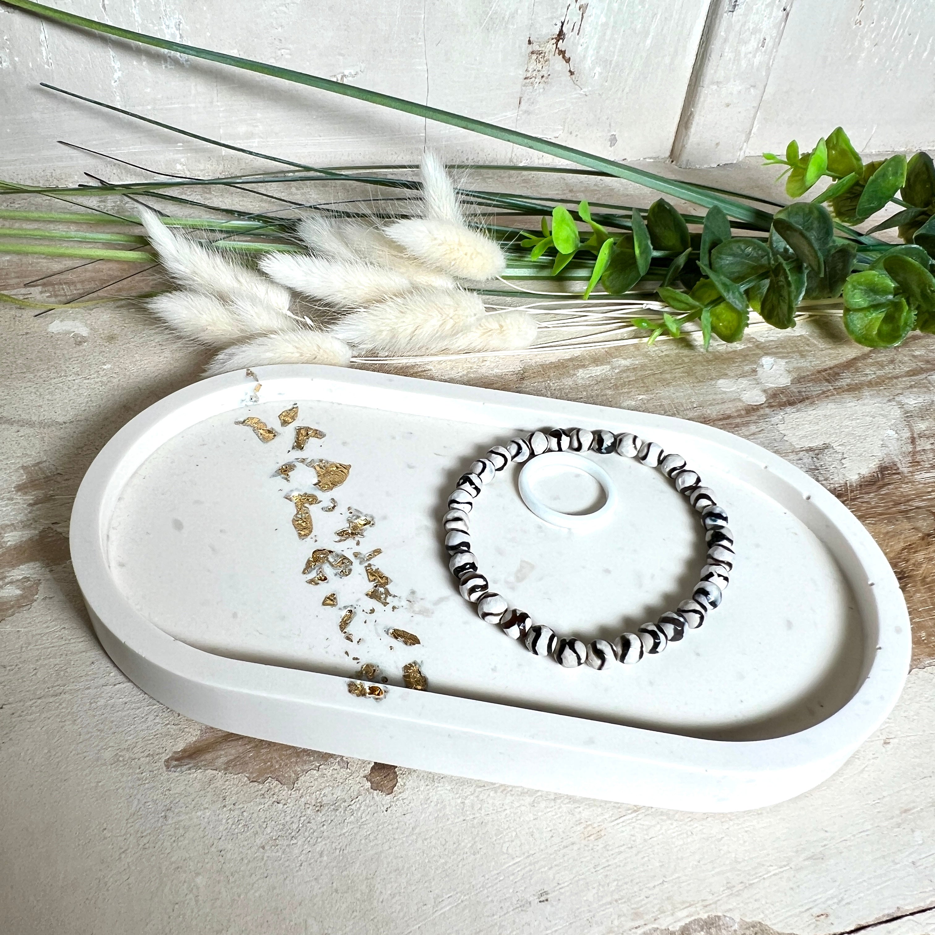 Handgefertigtes ovales Schmucktablett aus Jesmonite mit schimmernden Blattgold-Akzenten. Die Rückseite ist mit rutschfesten Korkpunkten ausgestattet. Ideal zur stilvollen Aufbewahrung von Schmuck, Schlüsseln oder kleinen Accessoires auf Kommoden und Nachttischen. Helles Design mit natürlicher Oberfläche und modernem Look. Unikat in feiner Handarbeit gefertigt – ohne Deko geliefert.