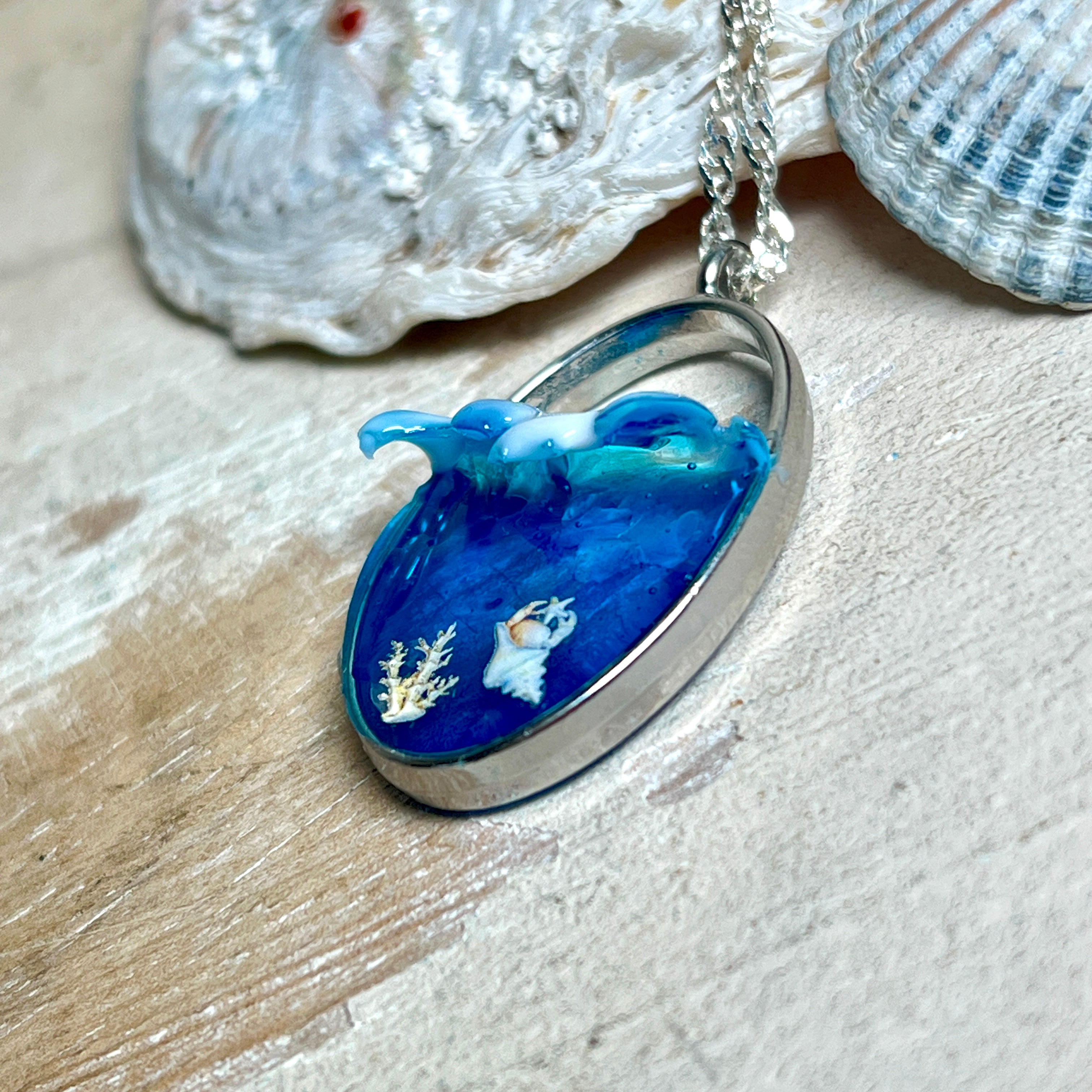 „Riffschatz“ – ovaler Harz-Anhänger mit plastischer 3D-Welle in Blau/Türkis, weißer Schaumkrone und Muschel-/Korallen-Stickern am Grund. Die Sticker sind unter klarem Harz versiegelt. Silber-Legierung, ca. 3,9 × 2,3 cm, an einer ca. 56 cm langen versilberten Kette. Handgefertigt, hochglänzend, jedes Stück ein Unikat.