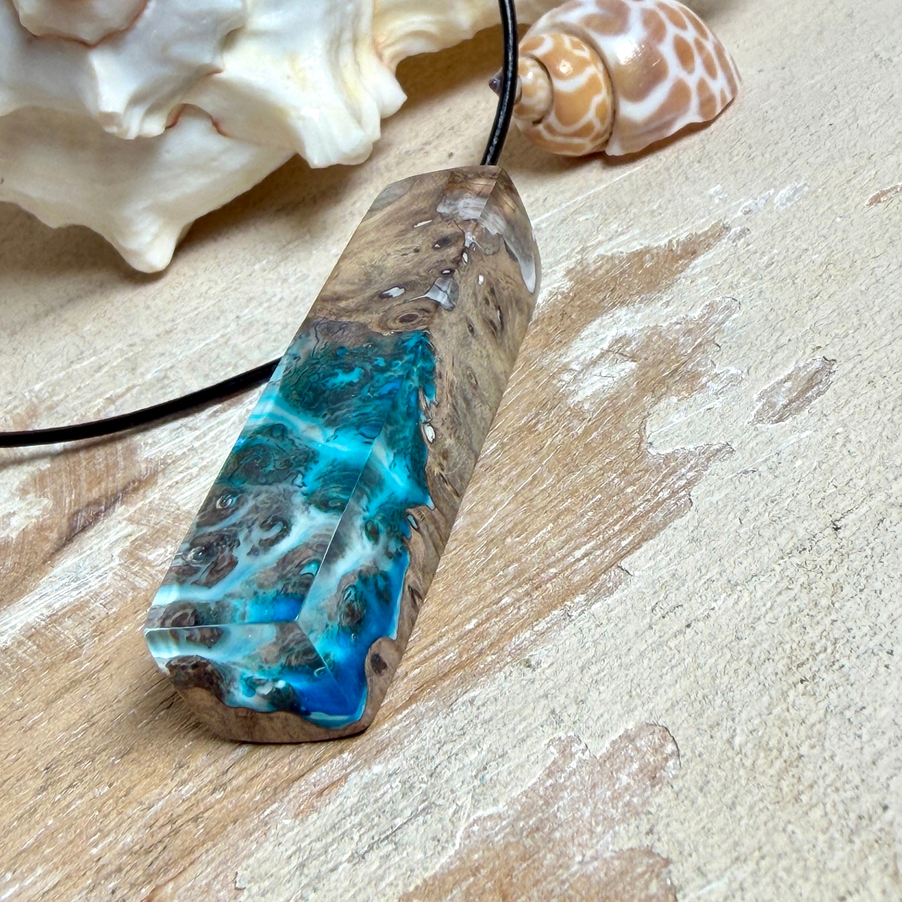 Rechteckiger Stabanhänger aus Maserholz und glasklarem Epoxidharz in tiefen Blau- und Türkistönen mit organischer Lagunen/Riff-Optik. Hochglanzpoliert, ca. 5,3 × 1,2 cm. Der Anhänger hängt an einer schwarzen Lederkette mit Edelstahl-Karabiner und 5-cm-Verlängerung (45 + 5 cm); die Anhängerschlaufe ist ebenfalls aus Edelstahl. Handgefertigtes Unikat, Lieferung in edler Schmuckbox – maritimer Look.