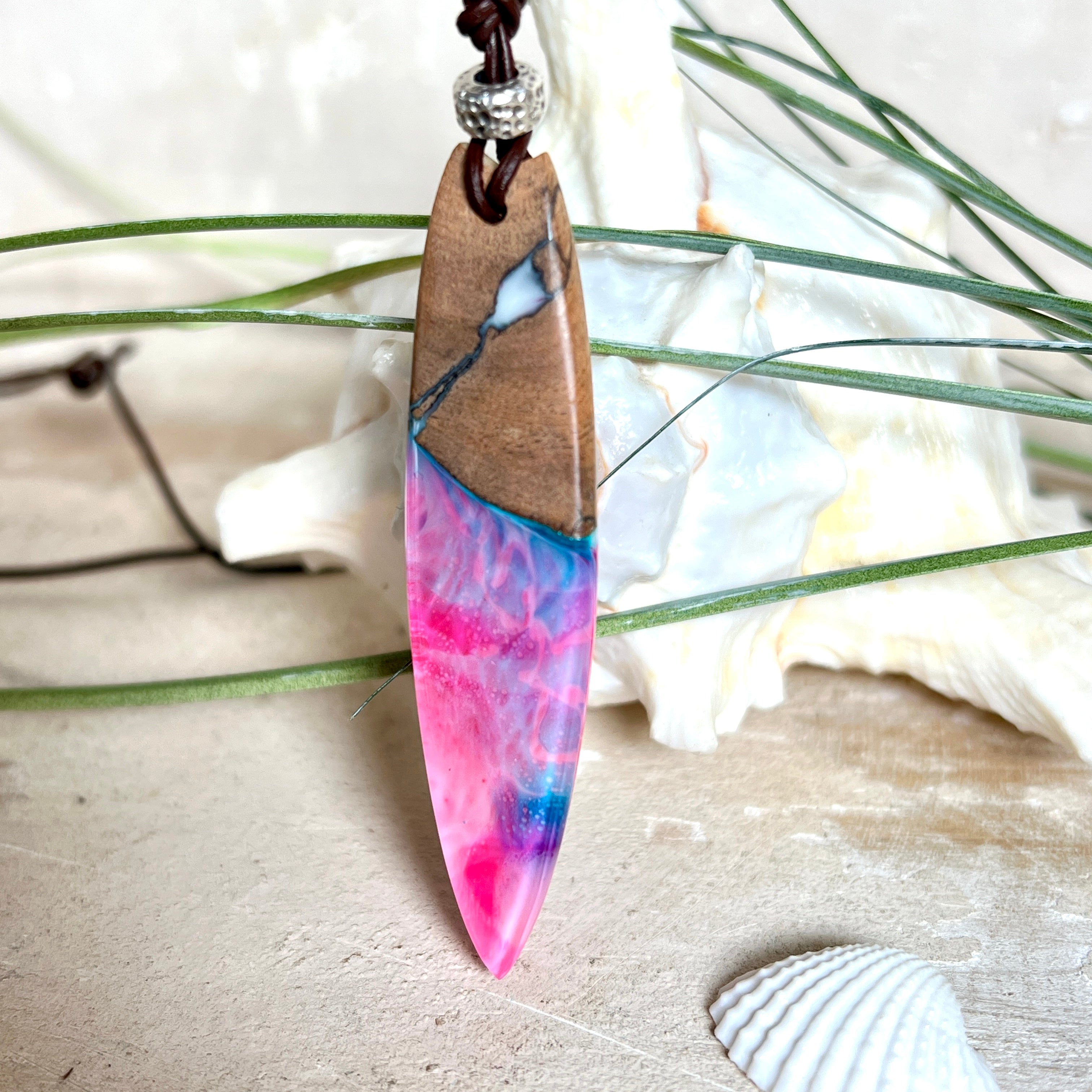 Handgefertigter Surfbrett-Anhänger aus Nussbaumholz und fluoreszierendem Epoxidharz in Pink, Blau und Weiß. Der Anhänger leuchtet zart bei Schwarzlicht und wird an einem verstellbaren Lederband (25–44 cm) mit Holzperlen getragen. Ein maritimes Unikat – ideal für Surfer und Naturliebhaber.