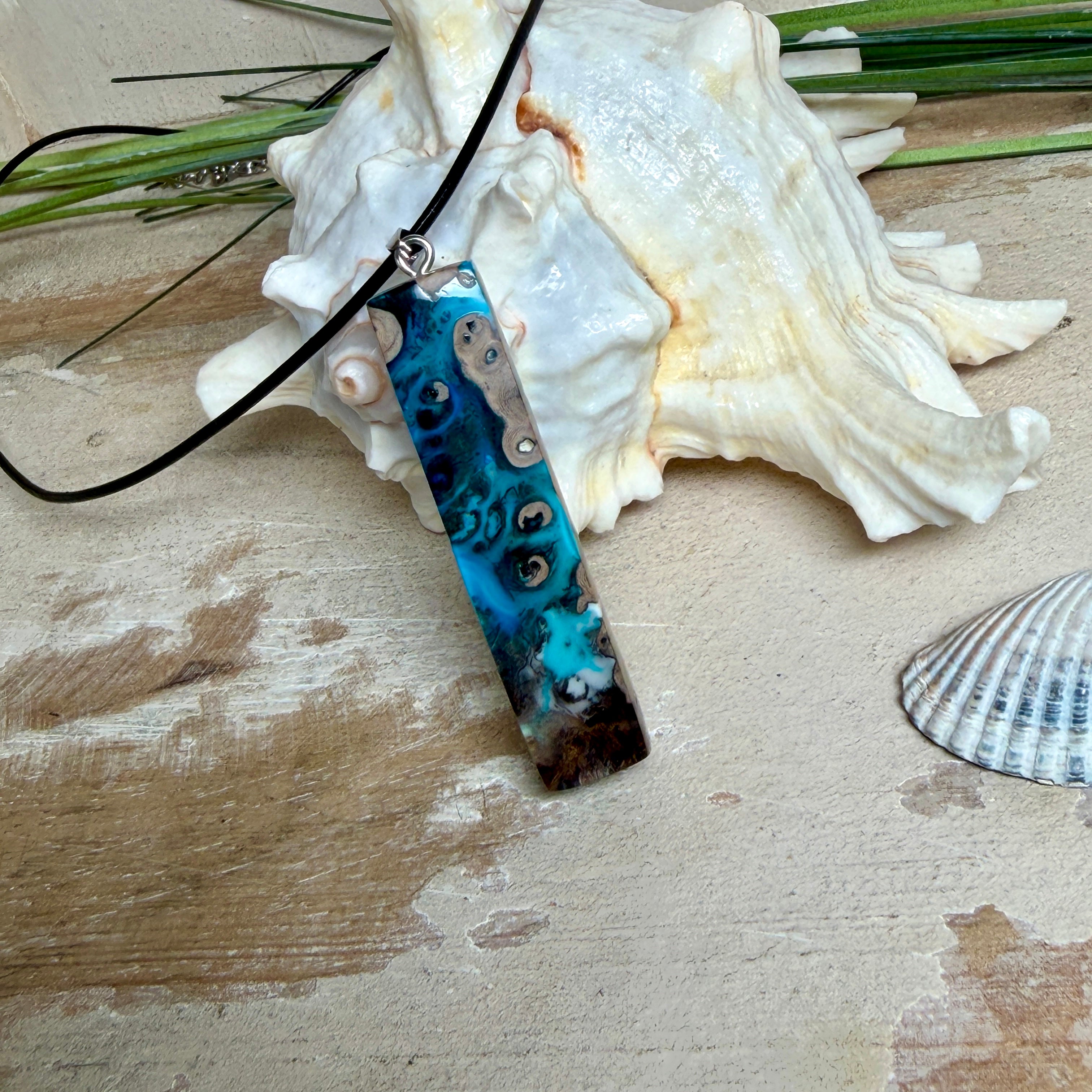 Rechteckiger Stabanhänger aus Maserholz und glasklarem Epoxidharz in tiefen Blau- und Türkistönen mit organischer Lagunen/Riff-Optik. Hochglanzpoliert, ca. 5,3 × 1,2 cm. Der Anhänger hängt an einer schwarzen Lederkette mit Edelstahl-Karabiner und 5-cm-Verlängerung (45 + 5 cm); die Anhängerschlaufe ist ebenfalls aus Edelstahl. Handgefertigtes Unikat, Lieferung in edler Schmuckbox – maritimer Look.