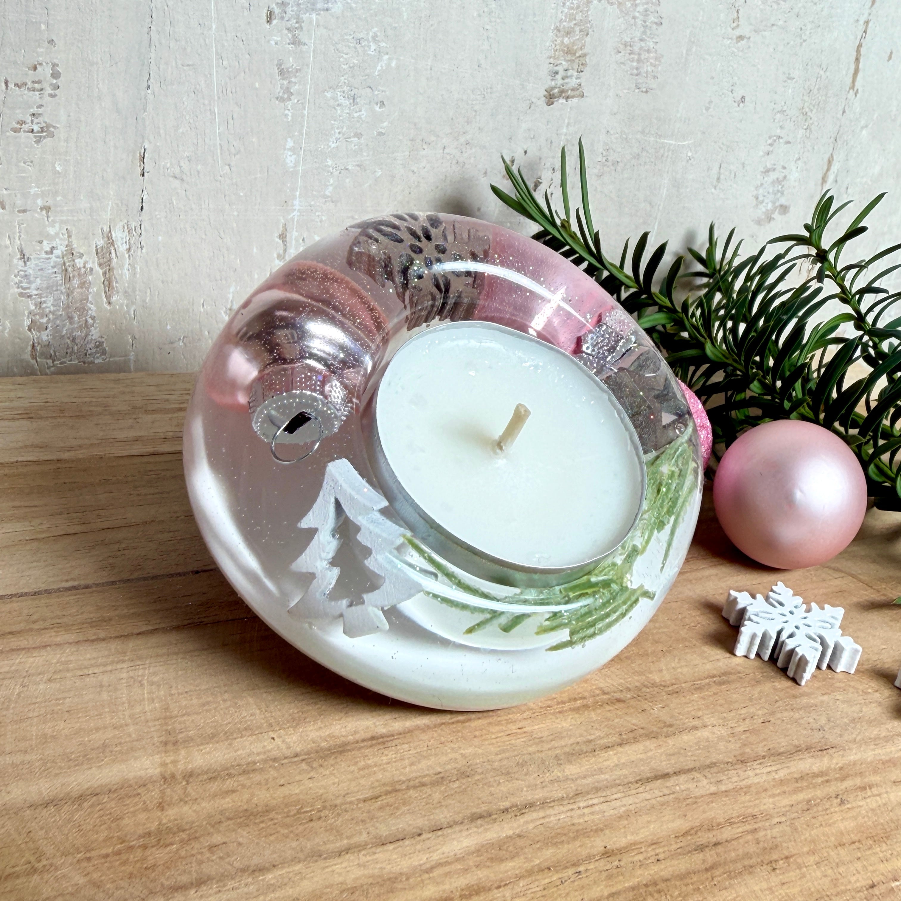 Runder Teelichthalter aus klarem Epoxidharz, Durchmesser ca. 7 cm, Höhe ca. 4 cm. Im Harz sind rosa Weihnachtskugeln, ein Stern, eine Schneeflocke, ein Tannenbaum, künstliche Tannennadeln und feiner Glitzer eingegossen. Vertiefung für ein Teelicht in der Mitte, Unterseite mit vier Korkpads als Möbelschoner. Fotografiert auf Holz mit Tannenzweig und rosa Kugeln als weihnachtliche Dekoration
