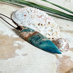 Unikat Anhänger aus stabilisiertem Holz mit türkisblauem Harzverlauf, schlanke Form, poliert. Verstellbares braunes Lederband 22–46 cm mit Schiebeknoten. Anhänger ca. 6,6 × 2 cm. Tragefoto am Hals und Produktfotos mit maritimer Deko