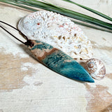 Unikat Anhänger aus stabilisiertem Holz mit türkisblauem Harzverlauf, schlanke Form, poliert. Verstellbares braunes Lederband 22–46 cm mit Schiebeknoten. Anhänger ca. 6,6 × 2 cm. Tragefoto am Hals und Produktfotos mit maritimer Deko