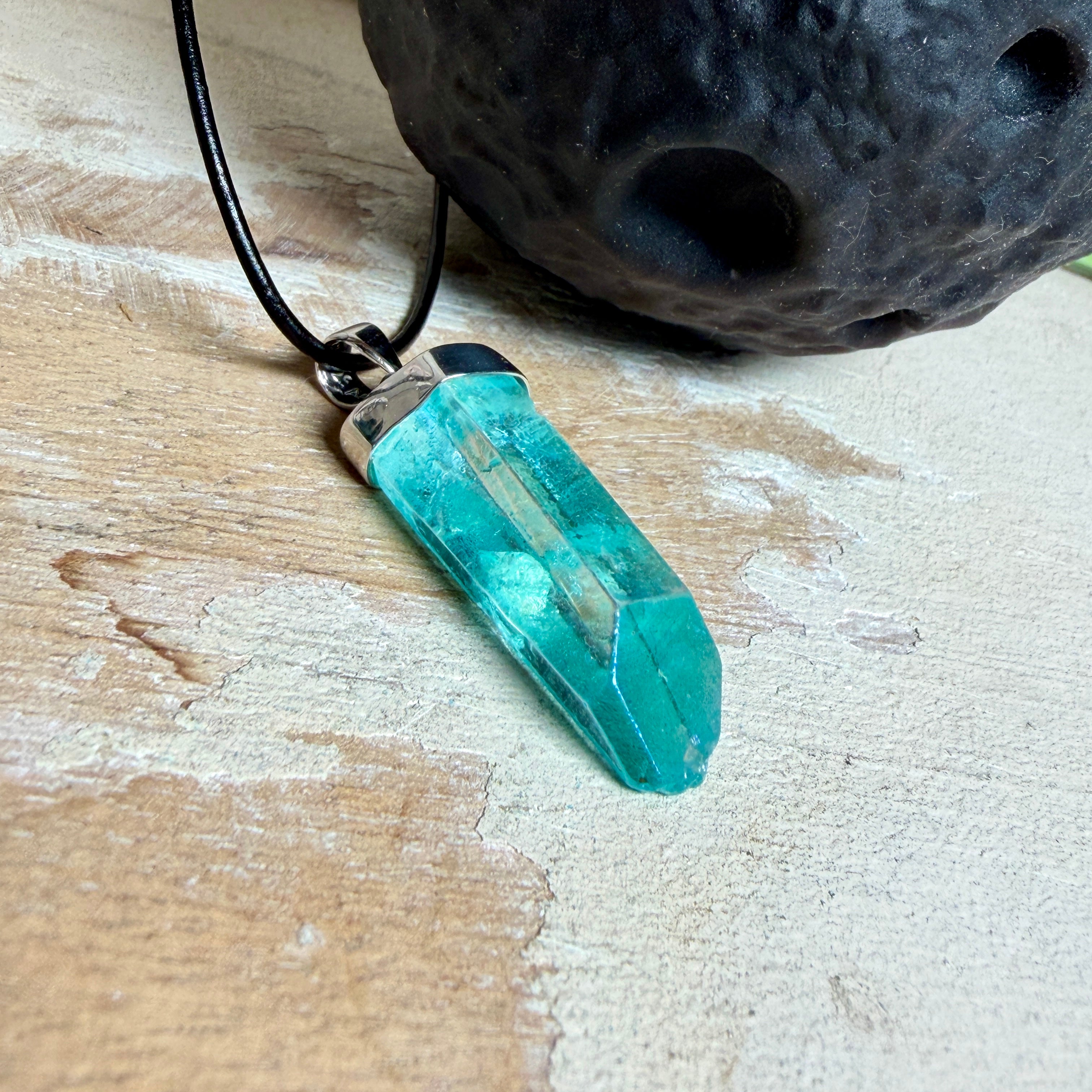Nahaufnahme einer handgefertigten Halskette mit Aqua Aura Rohkristall: naturbelassener, sechskantiger Bergkristall mit goldbedampfter Aqua-Beschichtung in Türkisblau, gefasst in 925er Silber und getragen an einer schwarzen Lederkette mit Edelstahl-Karabiner und Verlängerungskettchen.
