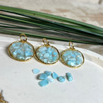 Runder Anhänger „Golden Atlantis Dream“ mit kleinen Larimar-Edelsteinchen in klarem Resin, gefasst in einer 24K vergoldeten Zinklegierung in Shiny Gold. Die Larimarsteine zeigen sanfte Blau- und Weißtöne wie karibische Wellen. Durchmesser ca. 22 mm, handgefertigtes Unikat, fotografiert mit Larimarsteinchen, Muscheln und einer 2-Euro-Münze zur Größenorientierung auf maritimem Untergrund.